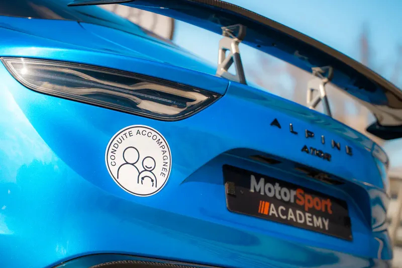 L'arrière d'une Alpine A110 R bleue portant un disque de conduite accompagnée et une plaque Motorsport Academy