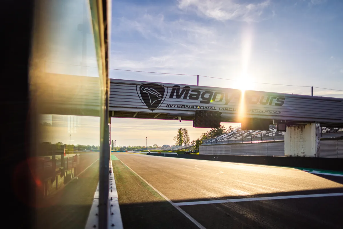 Ligne droite des stands sous la passerelle du circuit, à contre-jour au soleil levant