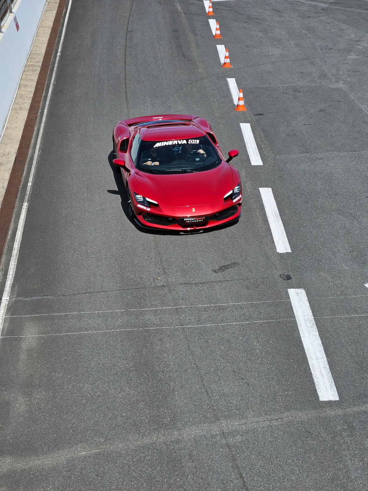 Une Ferrari 488 rouge aux couleurs Motorsport Academy, vue de dessus, à l'arrêt sur la voie des stands le long d'une file de cônes orange.